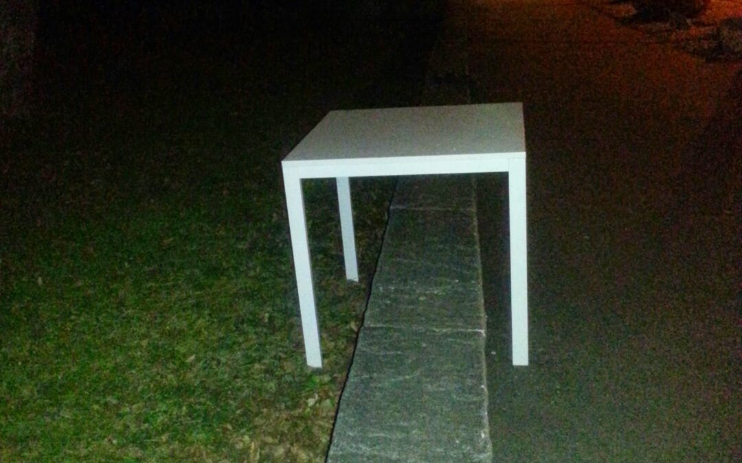Weisser Tisch bei Nacht, halb auf Gehweg, halb auf Rasen – symbolisiert fragile Balance und Zwischenräume demokratischer Zustände.